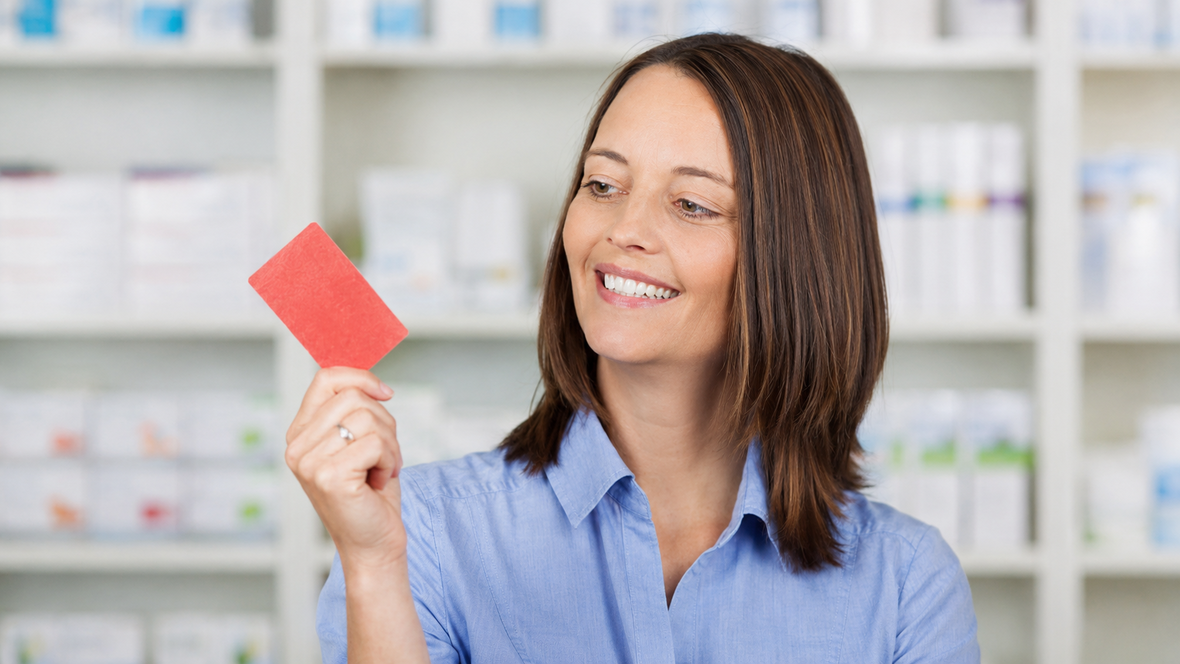 Eine lächelnde Kundin hält in einer Apotheke eine rote Kundenkarte in der Hand.