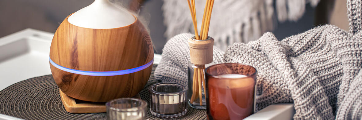 Aromatherapie mit einem dampfenden Duftdiffusor, Duftstäbchen und einer Kerze neben einer weichen Strickdecke.