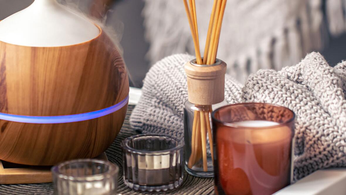 Aromatherapie mit einem dampfenden Duftdiffusor, Duftstäbchen und einer Kerze neben einer weichen Strickdecke.