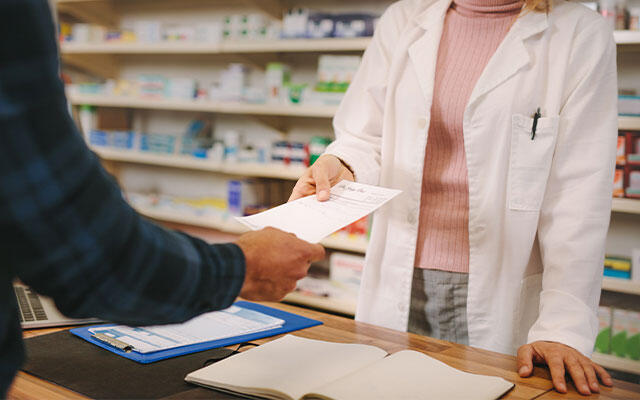 Kundin übergibt Rezept an Apothekerin in der Elisen-Apotheke. Arzneimittel im Hintergrund sichtbar.