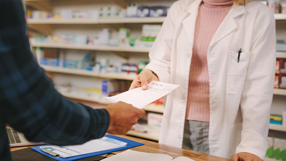 Kundin übergibt Rezept an Apothekerin in der Elisen-Apotheke. Arzneimittel im Hintergrund sichtbar.