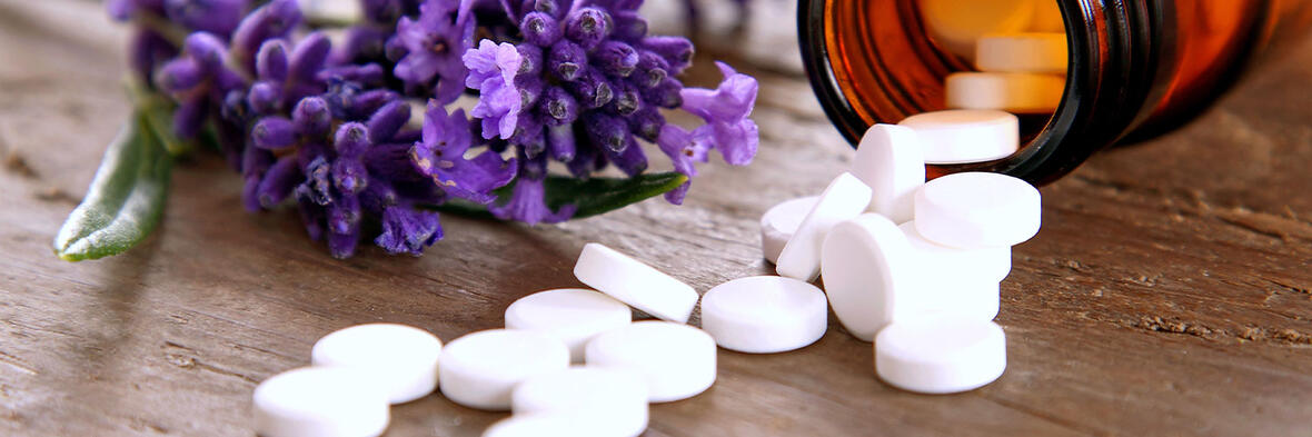 Weiße Schüßler-Salz-Tabletten liegen neben einer braunen Arzneimittelflasche und violetten Blüten auf einem Holztisch.