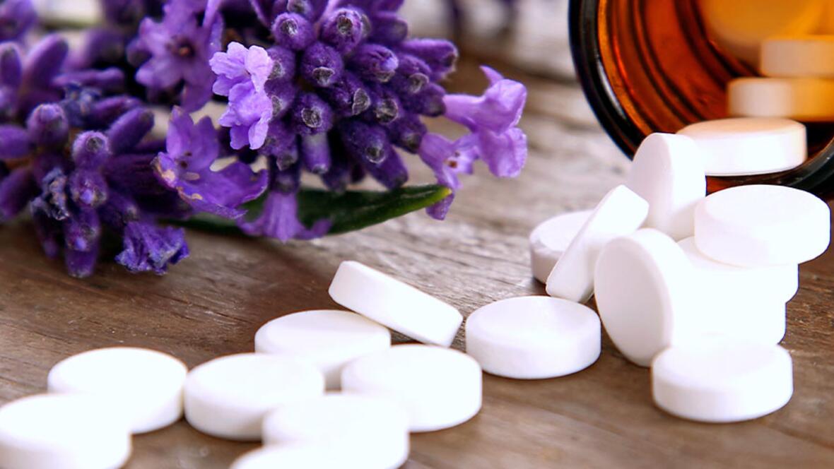 Weiße Schüßler-Salz-Tabletten liegen neben einer braunen Arzneimittelflasche und violetten Blüten auf einem Holztisch.