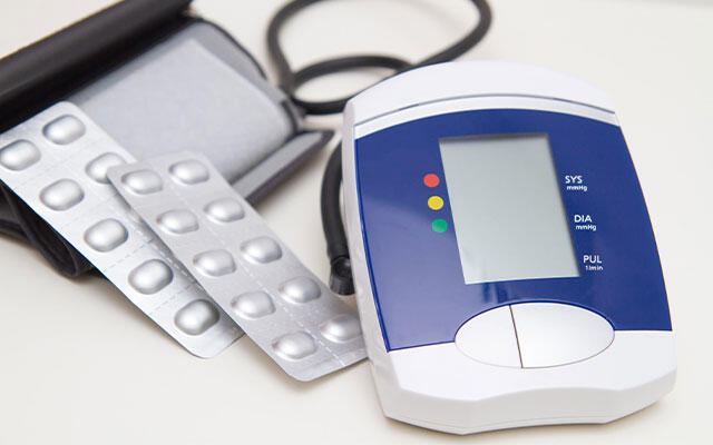 Digitales Blutdruckmessgerät mit Manschette neben mehreren Tablettenblistern.
