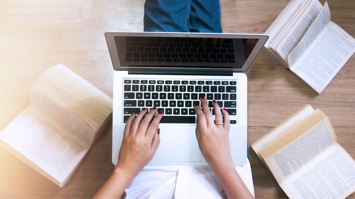 Hände tippen auf einer Laptop-Tastatur, umgeben von aufgeschlagenen Büchern auf einem Holztisch.