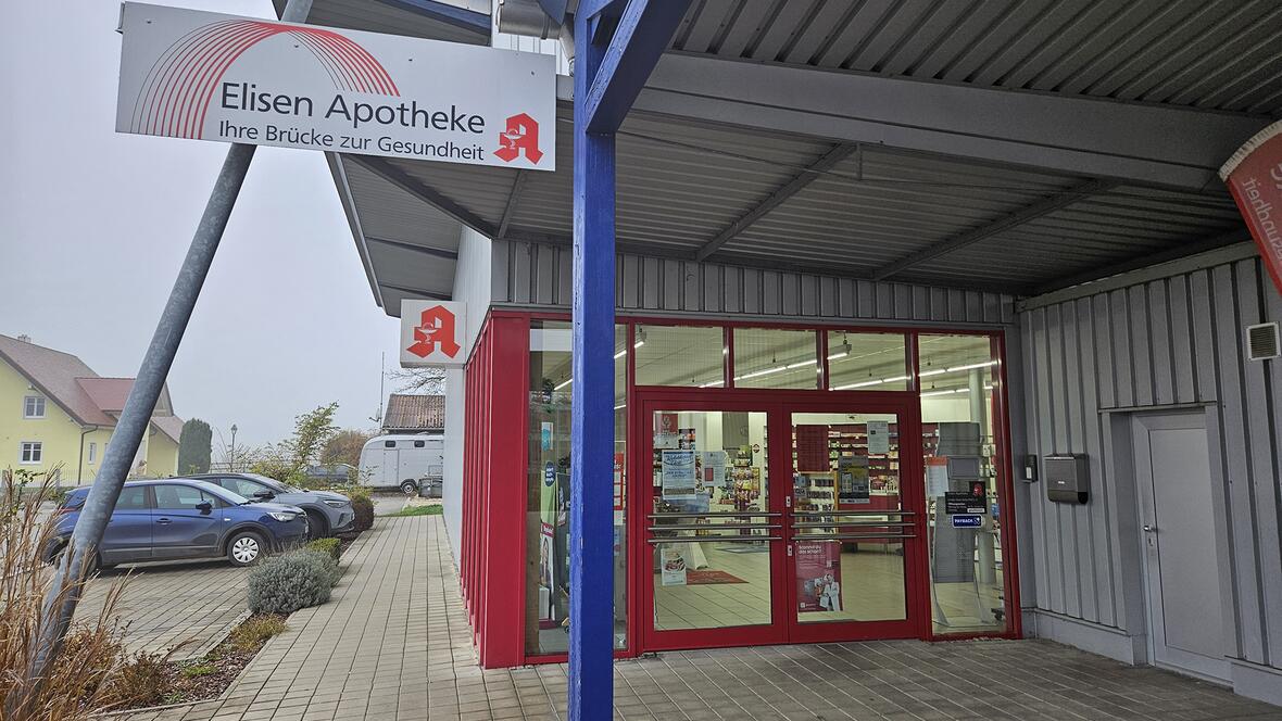 Eingang zur Elisen Apotheke mit roter Tür und Schildern, umgeben von Parkplatz und Nebel.
