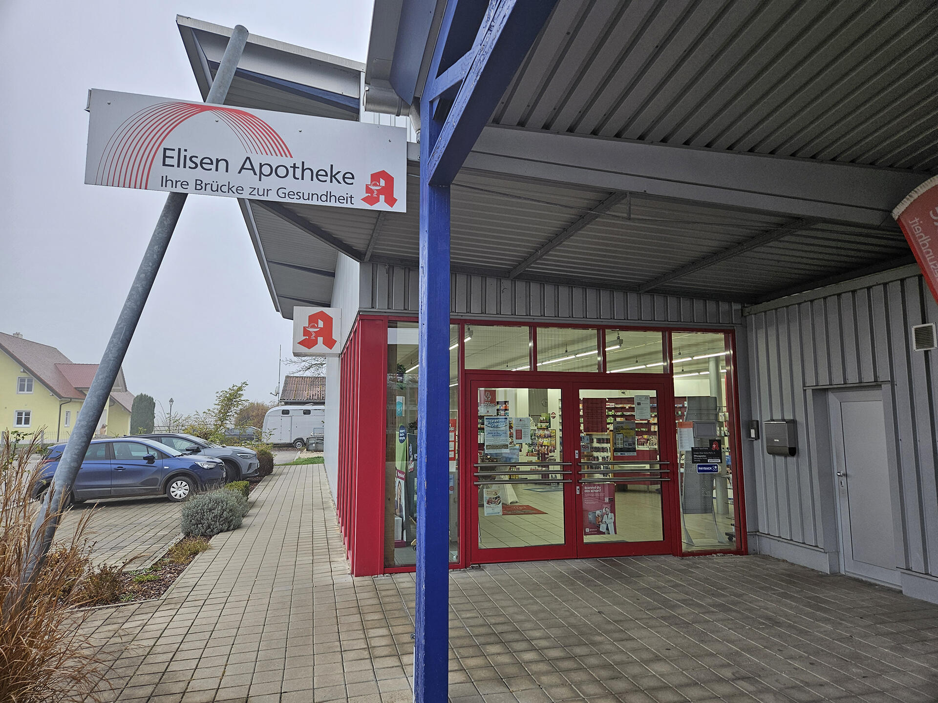 Eingang zur Elisen Apotheke mit roter Tür und Schildern, umgeben von Parkplatz und Nebel.