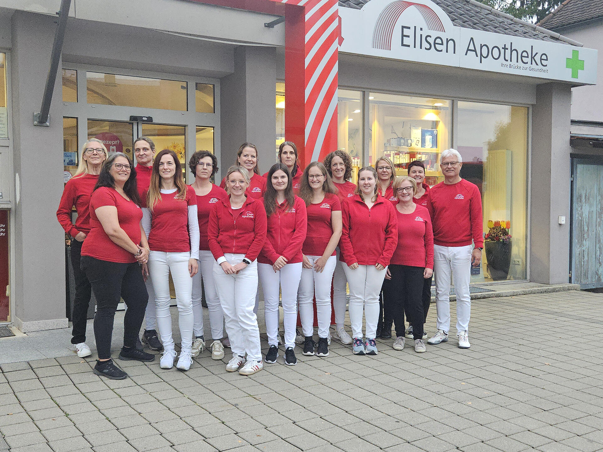 Mitarbeiterteam der Elisen Apotheke steht in roten Shirts vor dem Eingangsbereich.